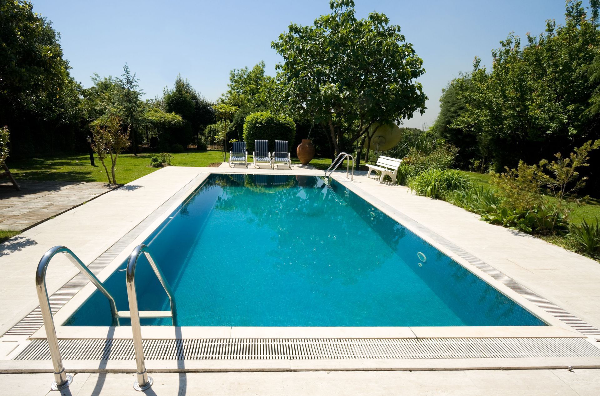 Outdoor backyard swimming pool with blue water, white deck, and lounge chairs surrounded by greenery