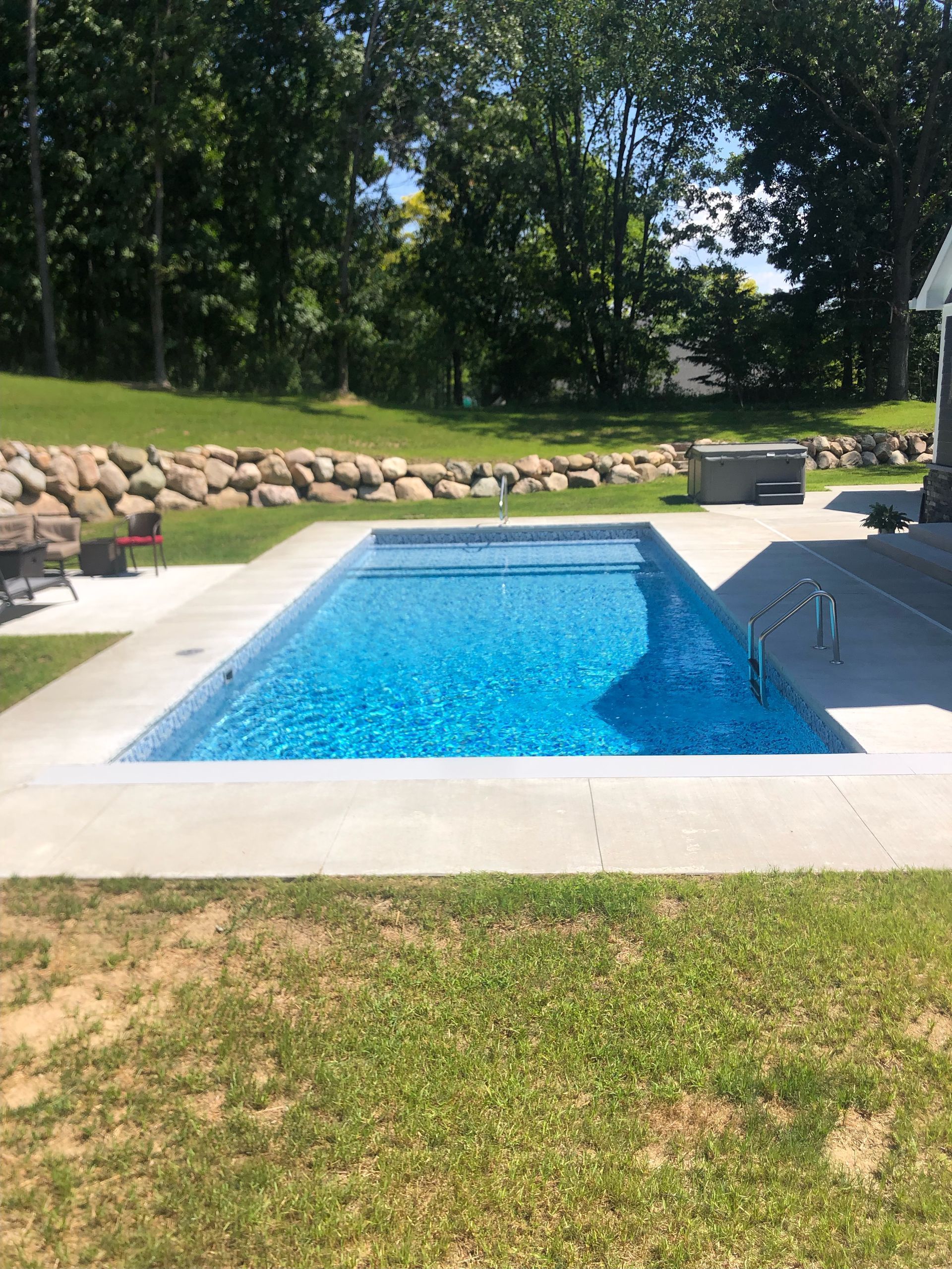 Backyard swimming pool with blue water, concrete deck, and a grassy lawn surrounded by trees.