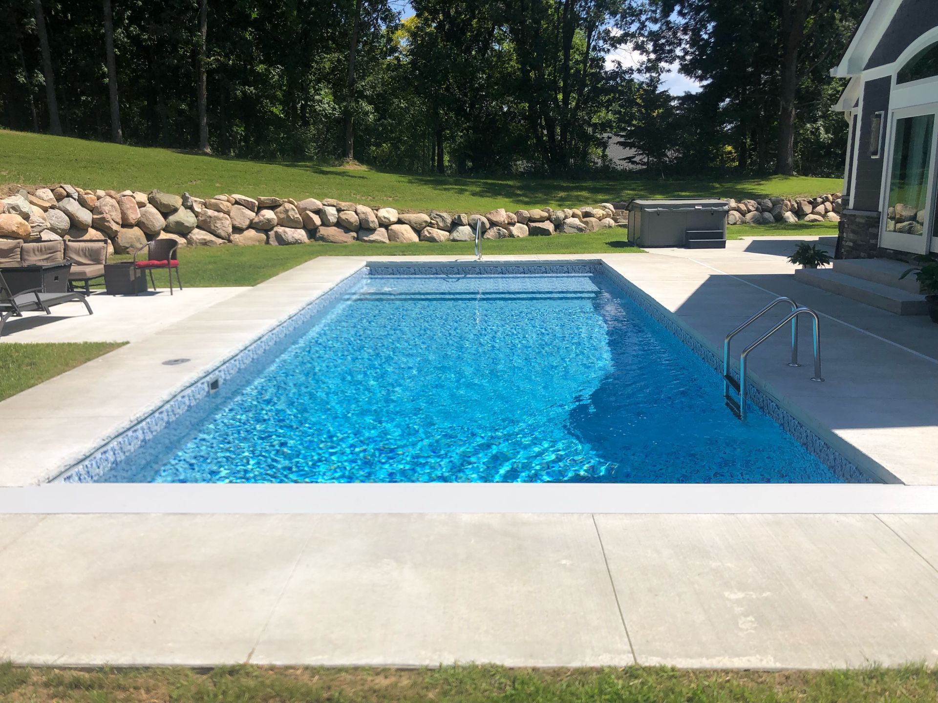 Backyard swimming pool with blue water, stone wall, and patio seating in sunlight