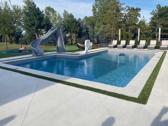 Outdoor pool with blue water, gray slide, and lounge chairs beside a concrete deck and trees