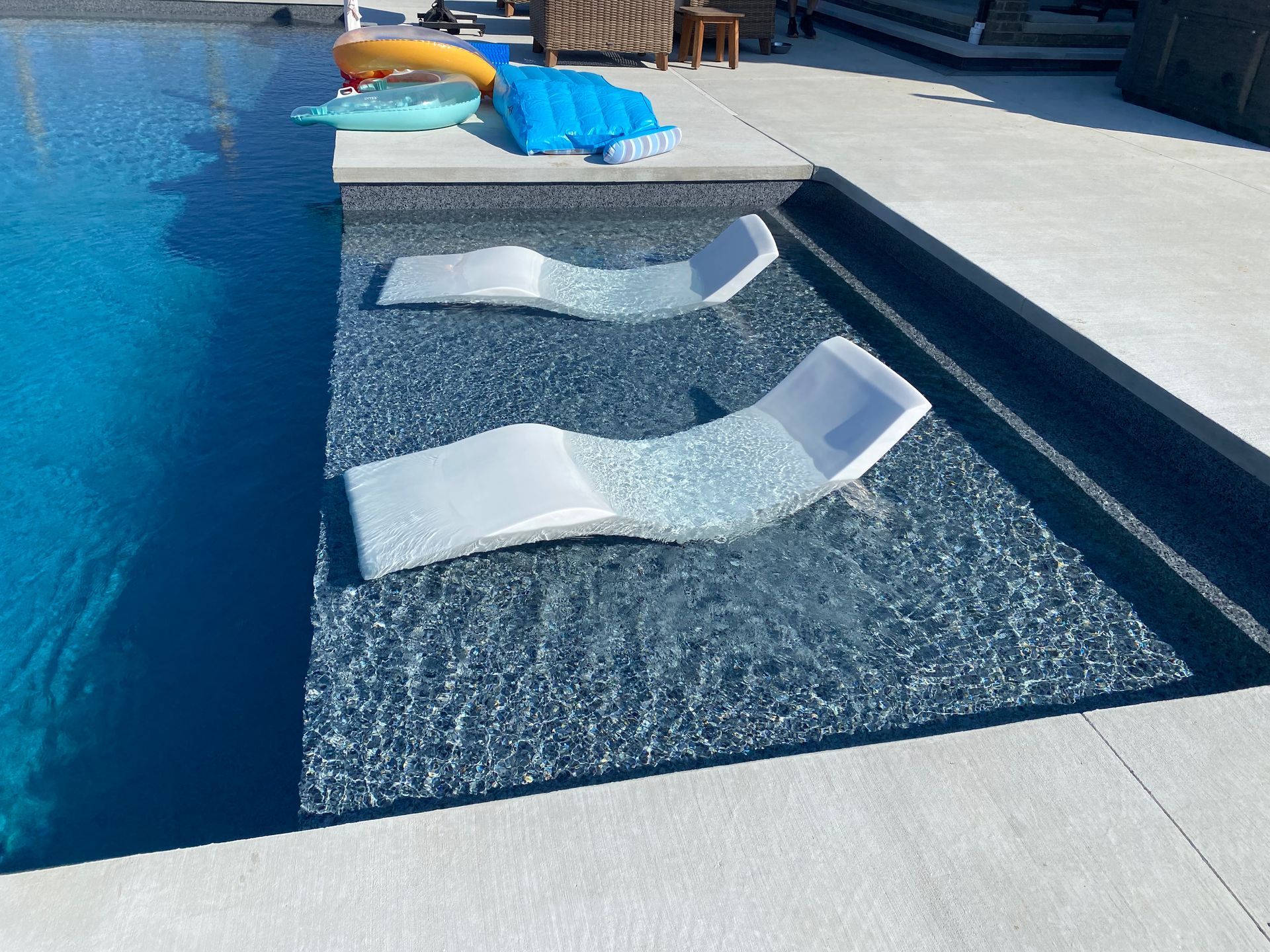 Poolside tanning ledge with two white in-water chaise lounges beside blue water