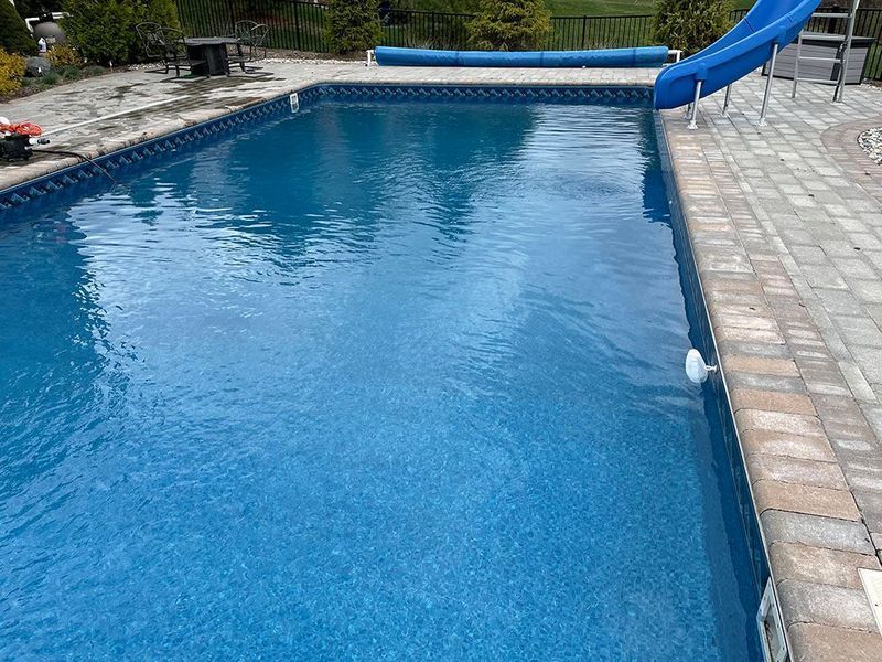 Rectangular swimming pool with blue water, surrounded by brick patio. A blue water slide is at one end.
