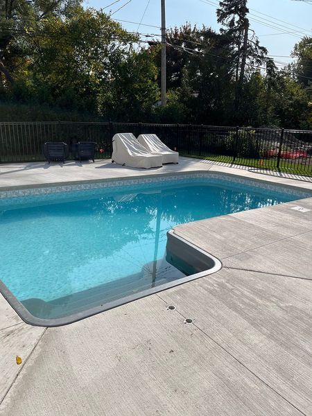 Swimming pool with concrete deck, slide, and black fence against a background of trees under a blue sky.
