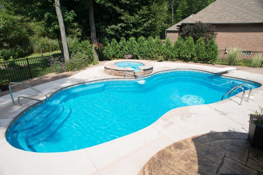 Blue, kidney-shaped swimming pool with attached hot tub, surrounded by a stone patio and landscaping.