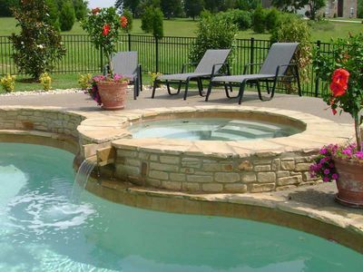 Pool with waterfall feature cascading into the pool, hot tub, lounge chairs, and potted flowers.