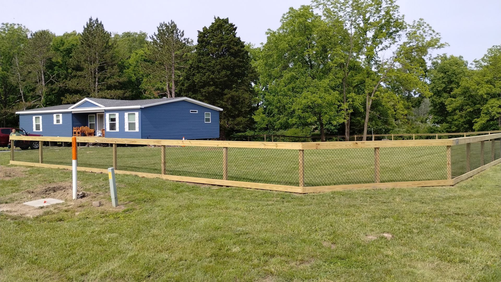 A blue manufactured home stands in a grassy yard enclosed by a wooden fence with wire mesh, surrounded by lush green trees.