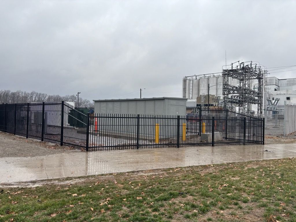 Industrial electrical site with fenced equipment, gray utility building, and cloudy sky near a grassy roadside