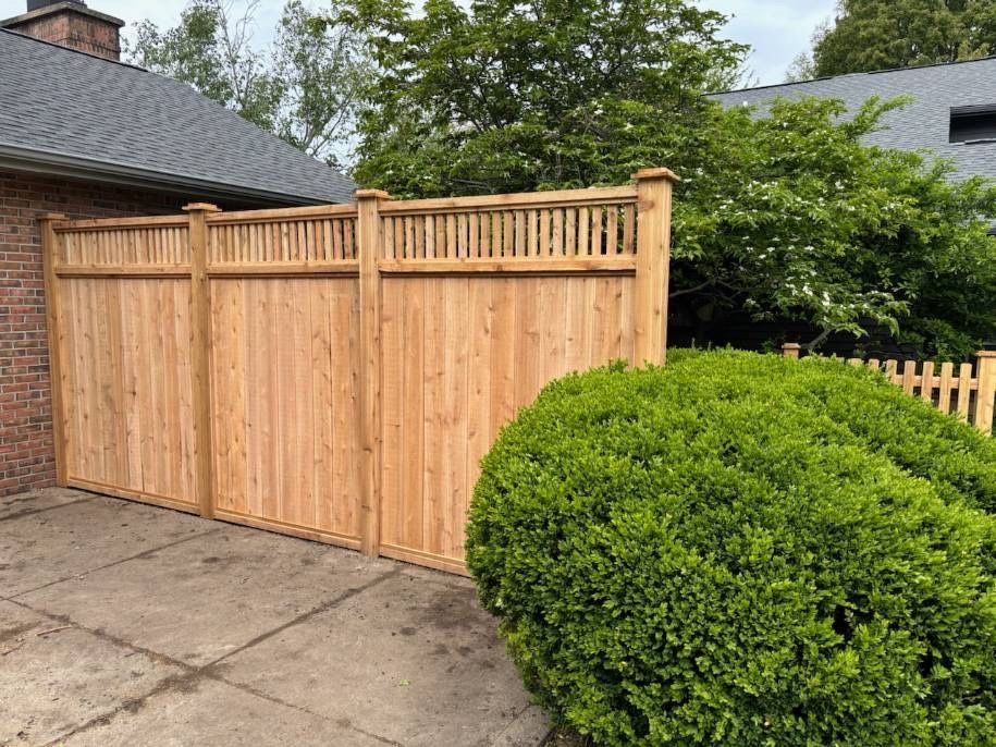 Wooden driveway gate beside a brick house, with a green hedge and trees in the background