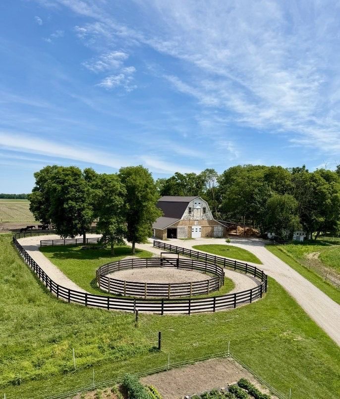 Farm scene with a fenced circular corral, white barn, trees, and a curving dirt road under a blue sky