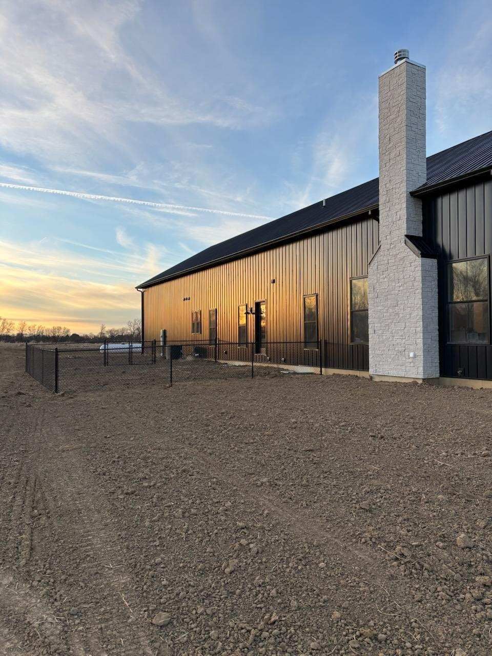 Large black building with a tall brick chimney, on a dirt lot, lit by a sunset.