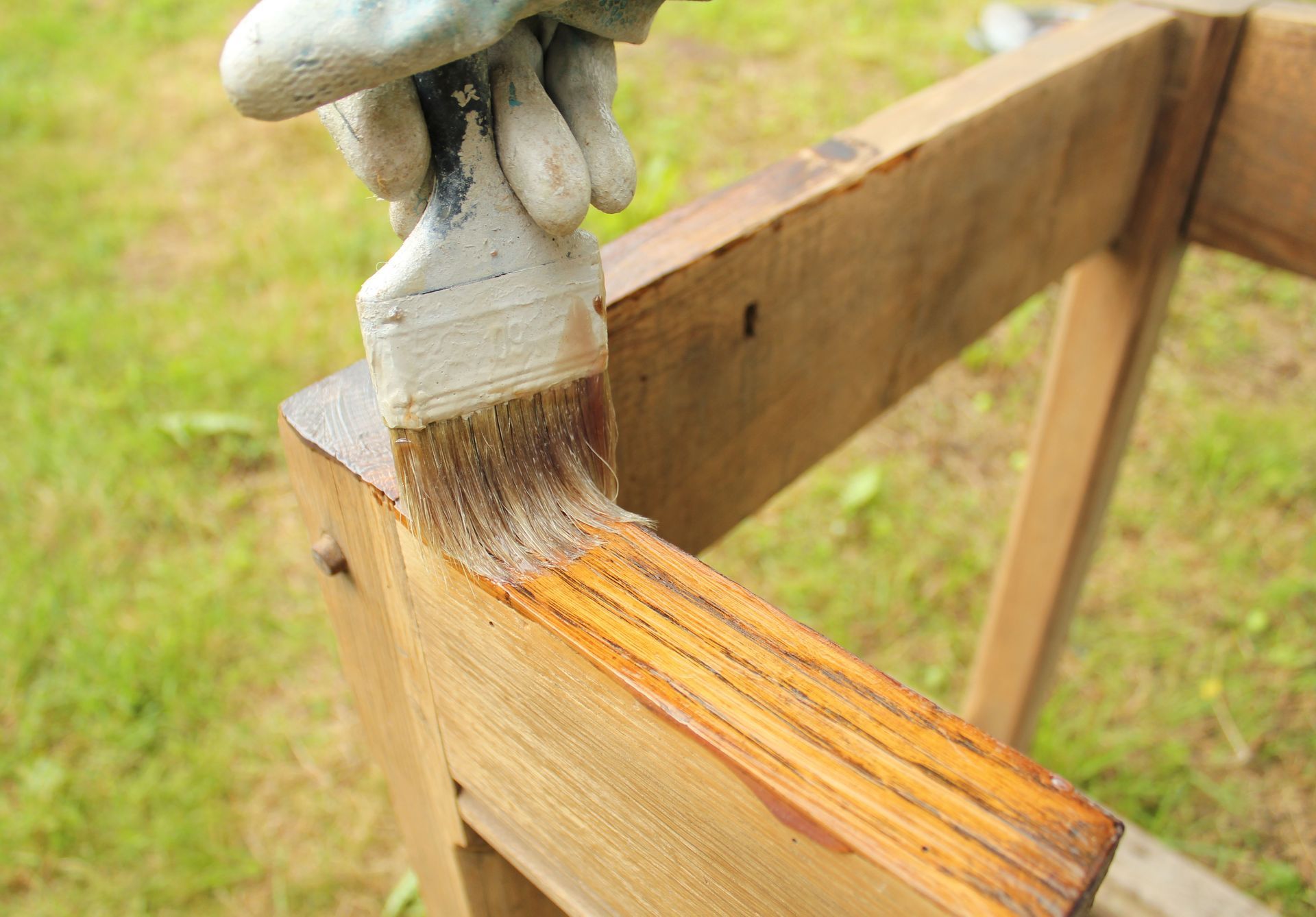 Person wearing a glove staining a wooden beam with a brush, outdoors.