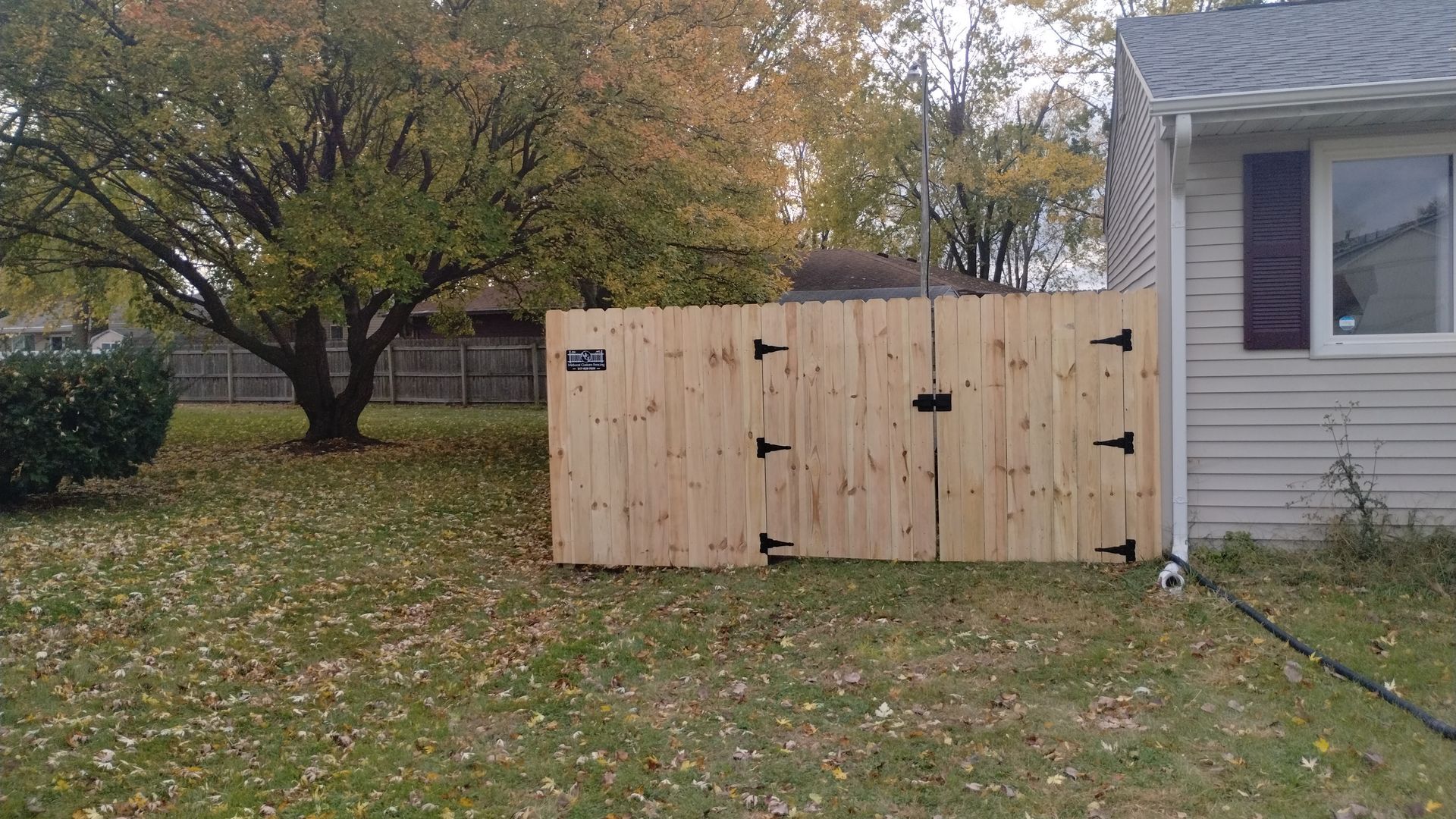 A light wood double gate stands in a grassy yard next to the side of a house with grey siding.