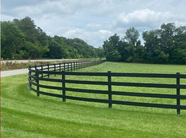 A black, three-rail wooden fence curves along a mowed green pasture beside a country road under a partly cloudy sky.