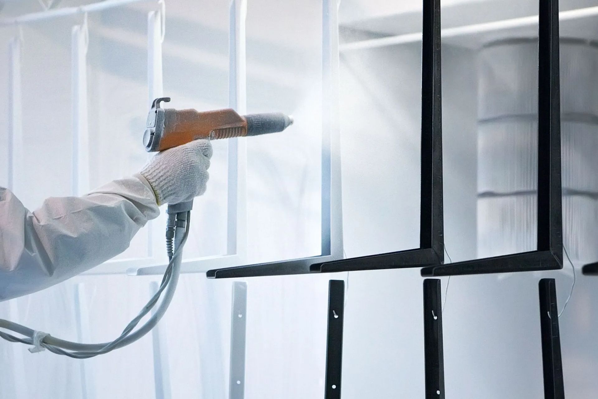 Person powder coating metal frame with orange spray gun in a white booth.