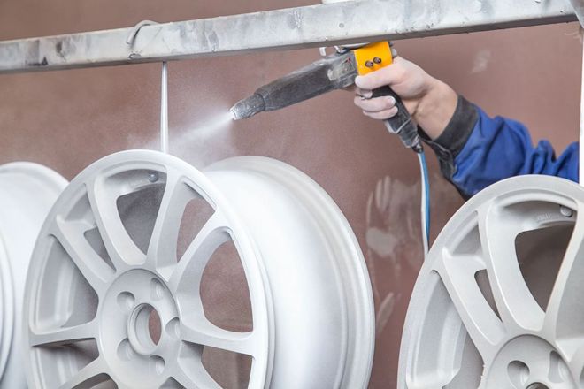 Person powder coating a set of white car rims with a spray gun, in a workshop.