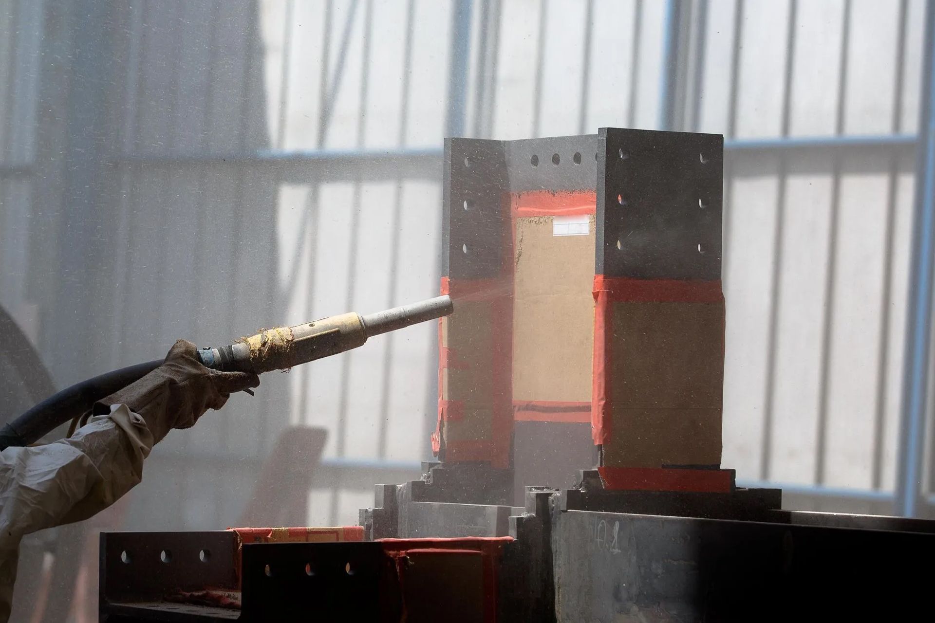 Sandblasting a rectangular metal frame in a workshop, creating a cloud of particles.