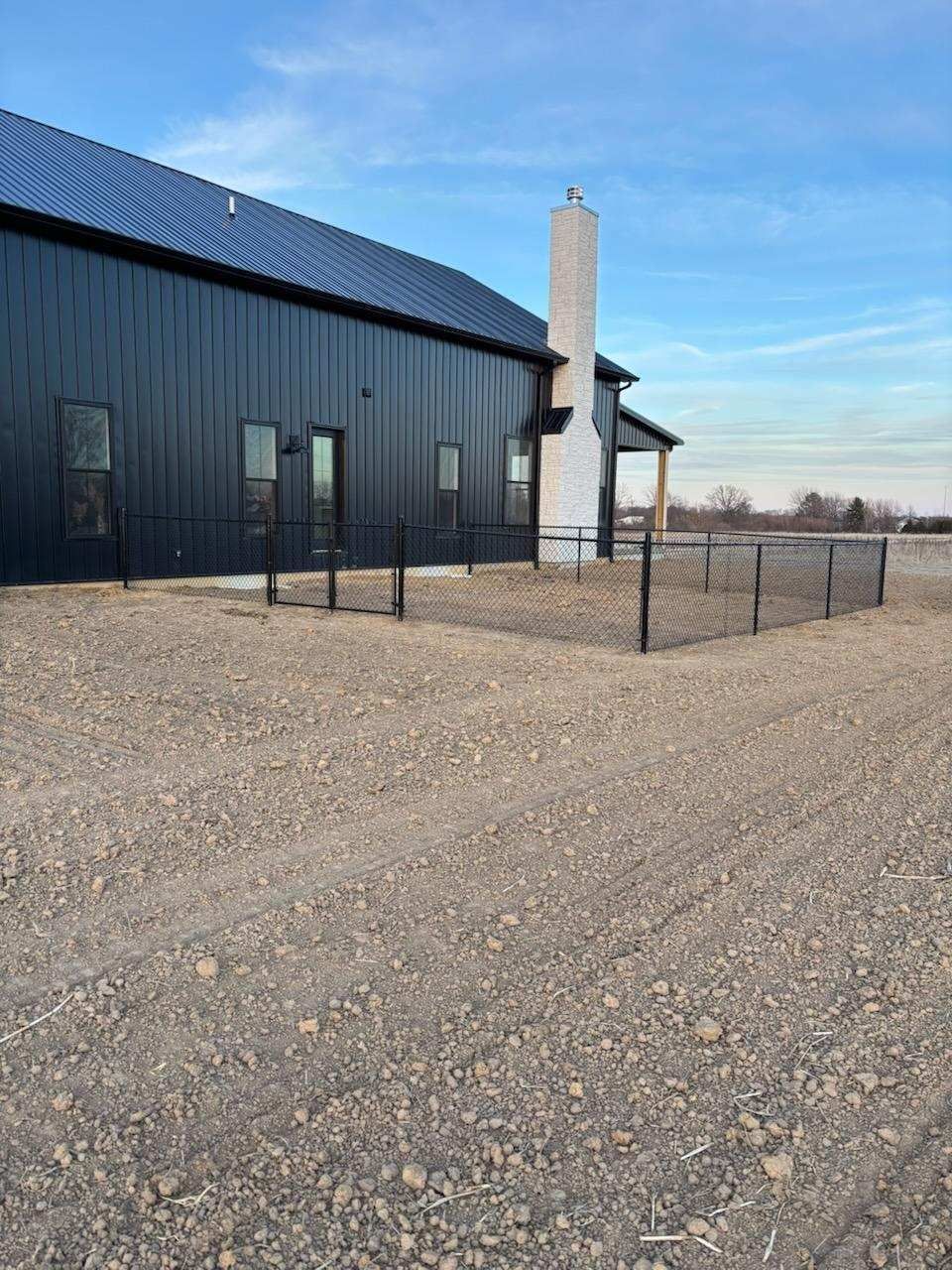 Black-sided modern barn with white chimney, small black fence, and gravel lot under a blue sky.