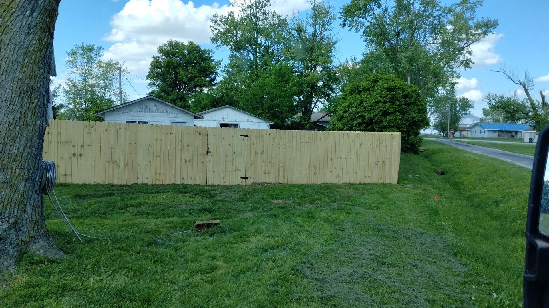 A new light-colored wood privacy fence stands in a grassy yard in front of a house on a sunny day.