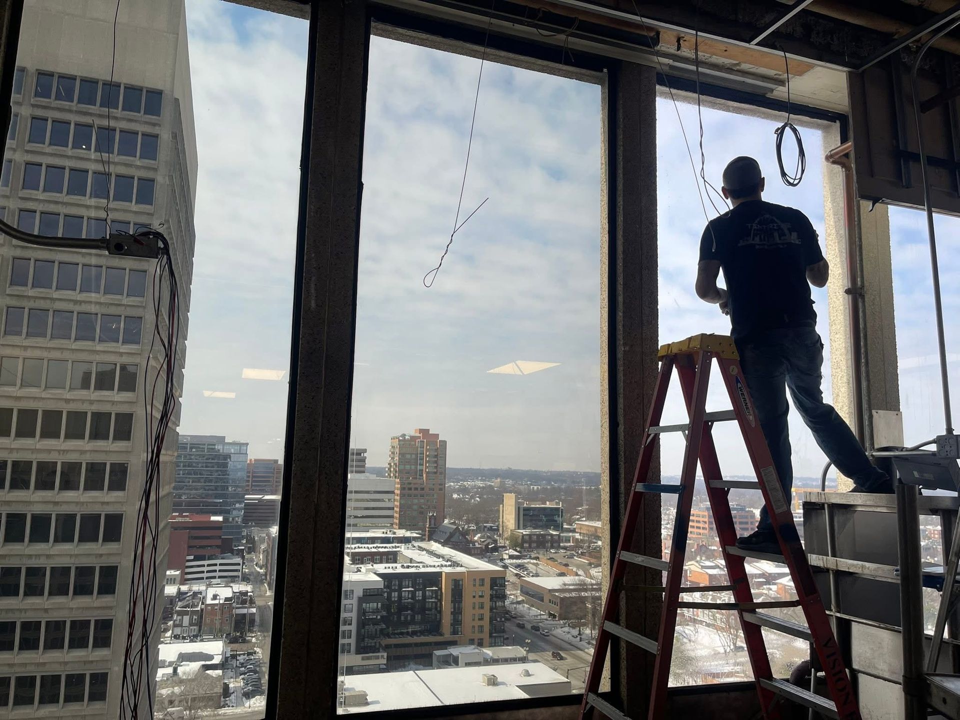 Worker on a ladder installing something near a large office window with a city view beyond.