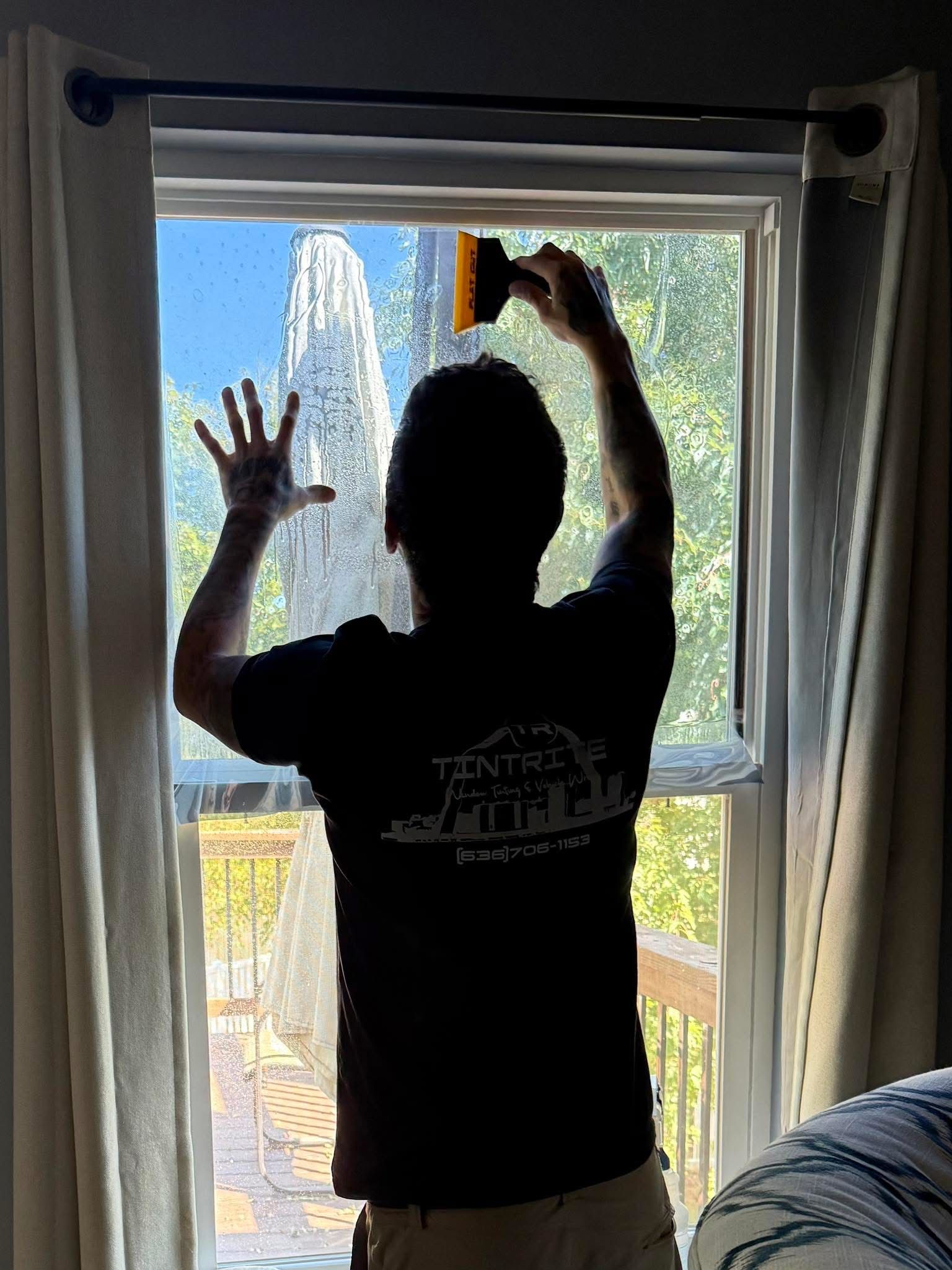 Silhouette of a person adjusting a window screen, with a blue sky and garden view outside.