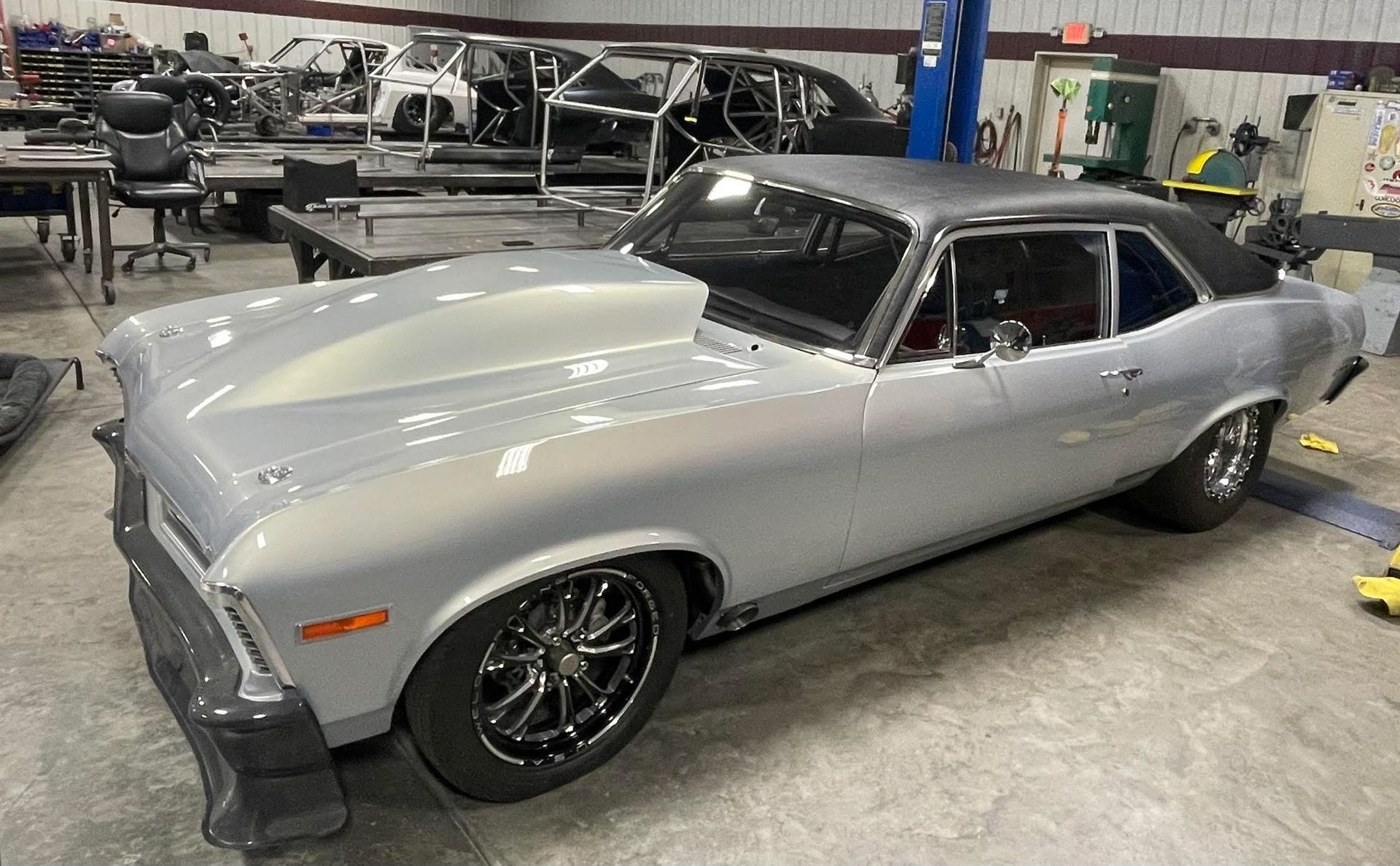 Silver classic coupe in an auto shop, with black wheels and hood stripes, parked indoors.
