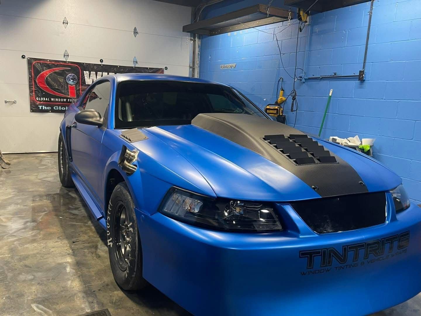 Blue sports car in a garage, front-left view, with a black hood vent and tinted windows.
