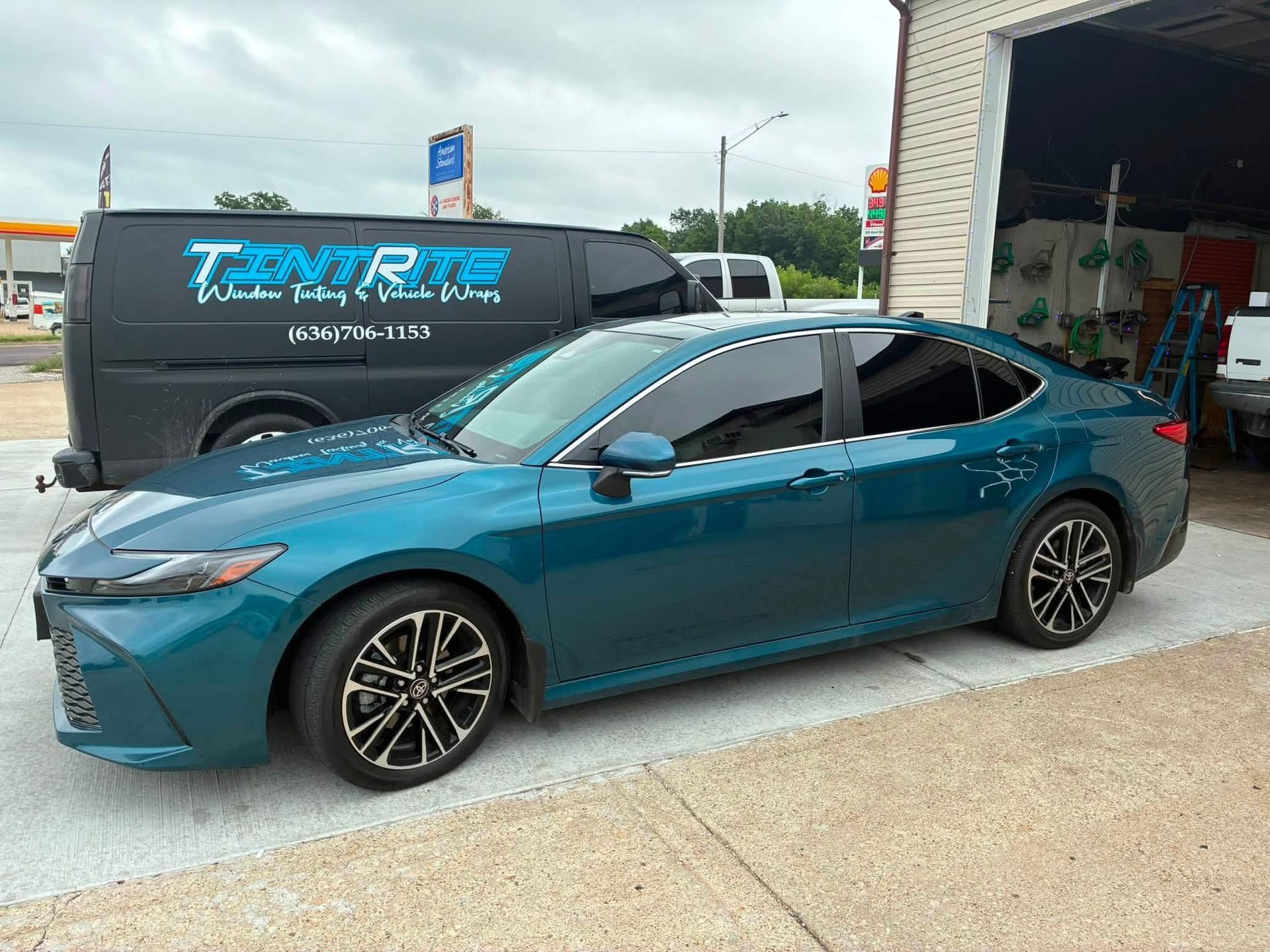 Blue sedan parked outside an auto shop with a black service van in the background.