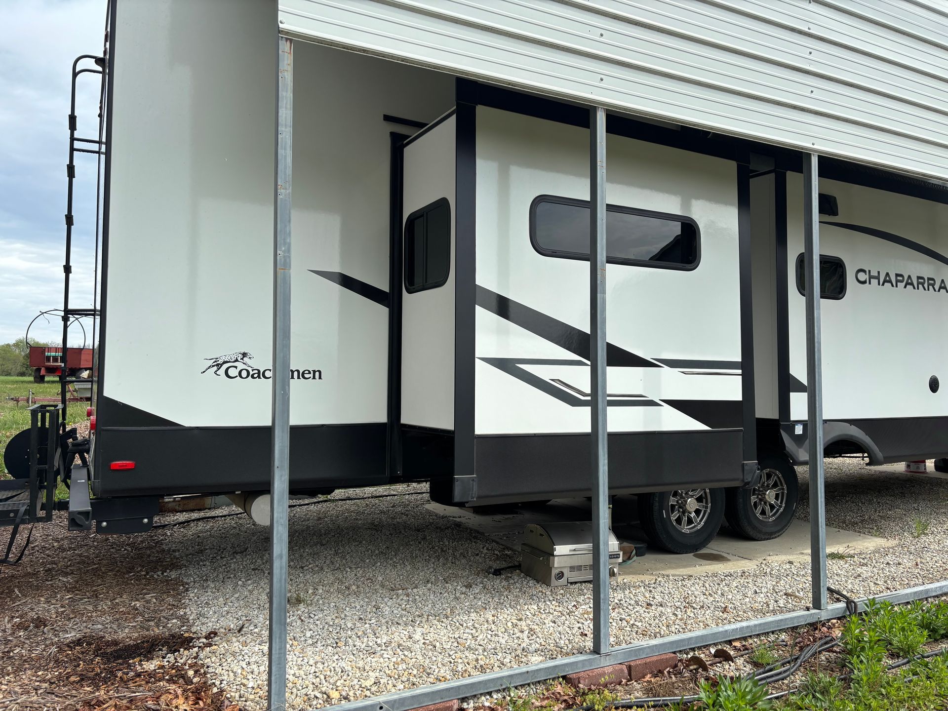White travel trailer with slide-out parked on gravel lot, viewed through metal posts.