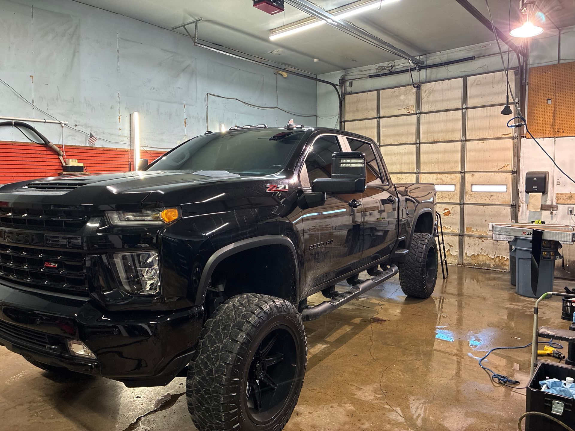 Black lifted pickup truck in a garage, wet floor reflecting overhead lights.