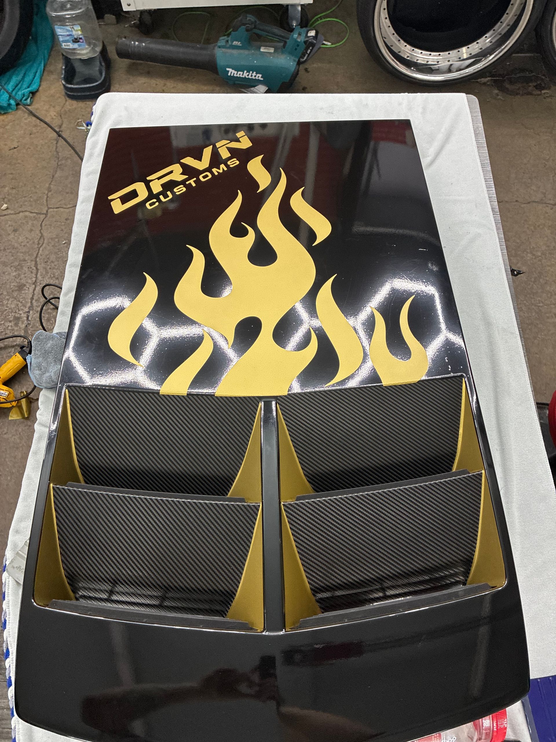 Black and gold custom car hood with flame graphics and four carbon-fiber panels on a workshop floor