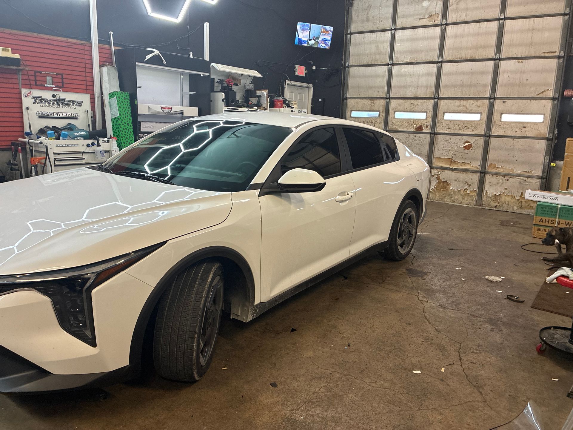 White sedan parked inside an auto shop garage, with a dusty floor and stacked cardboard boxes nearby