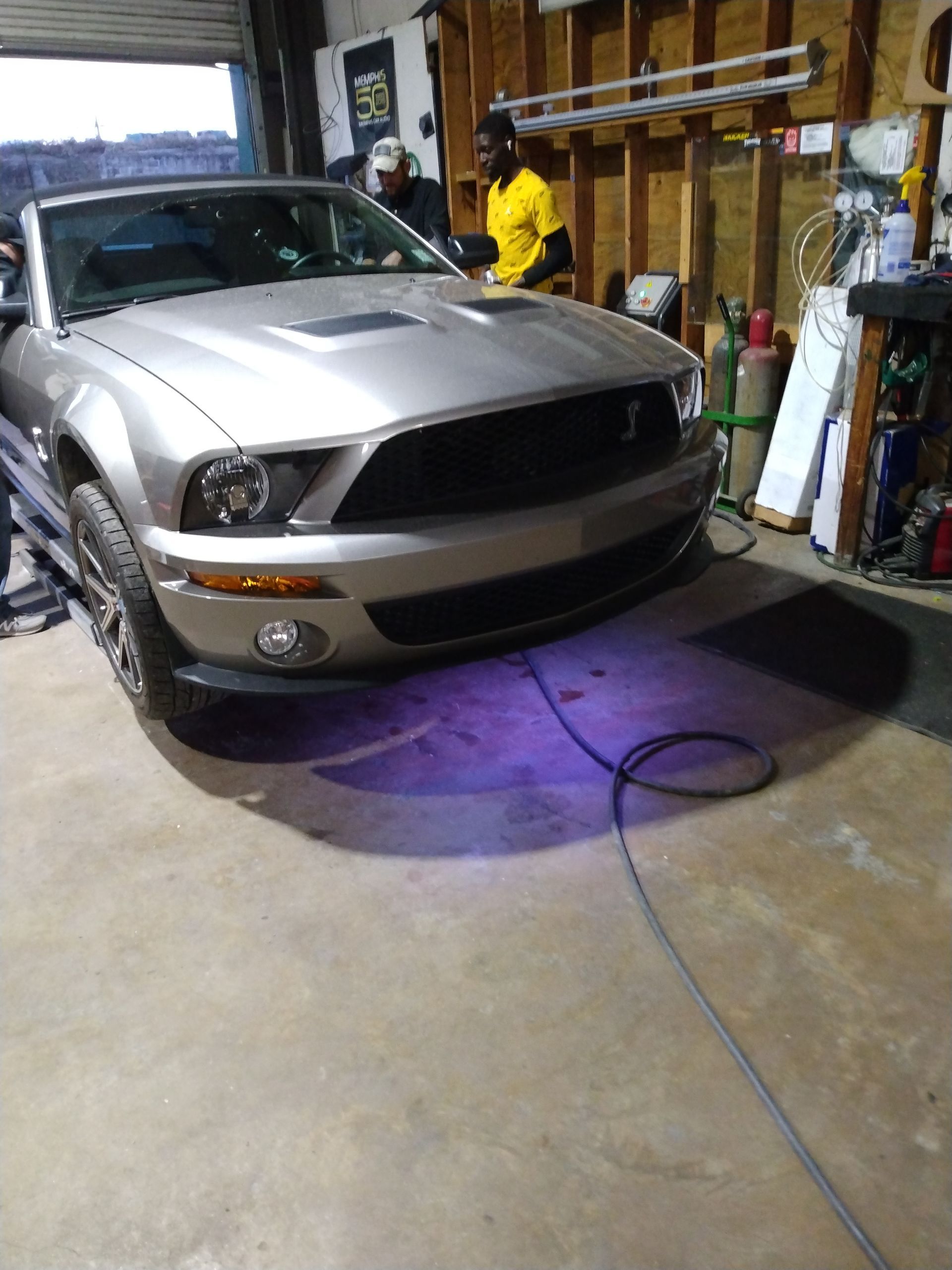 A silver mustang is parked in a garage with purple lights on the ground.