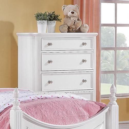 A teddy bear sits on top of a white dresser next to a bed
