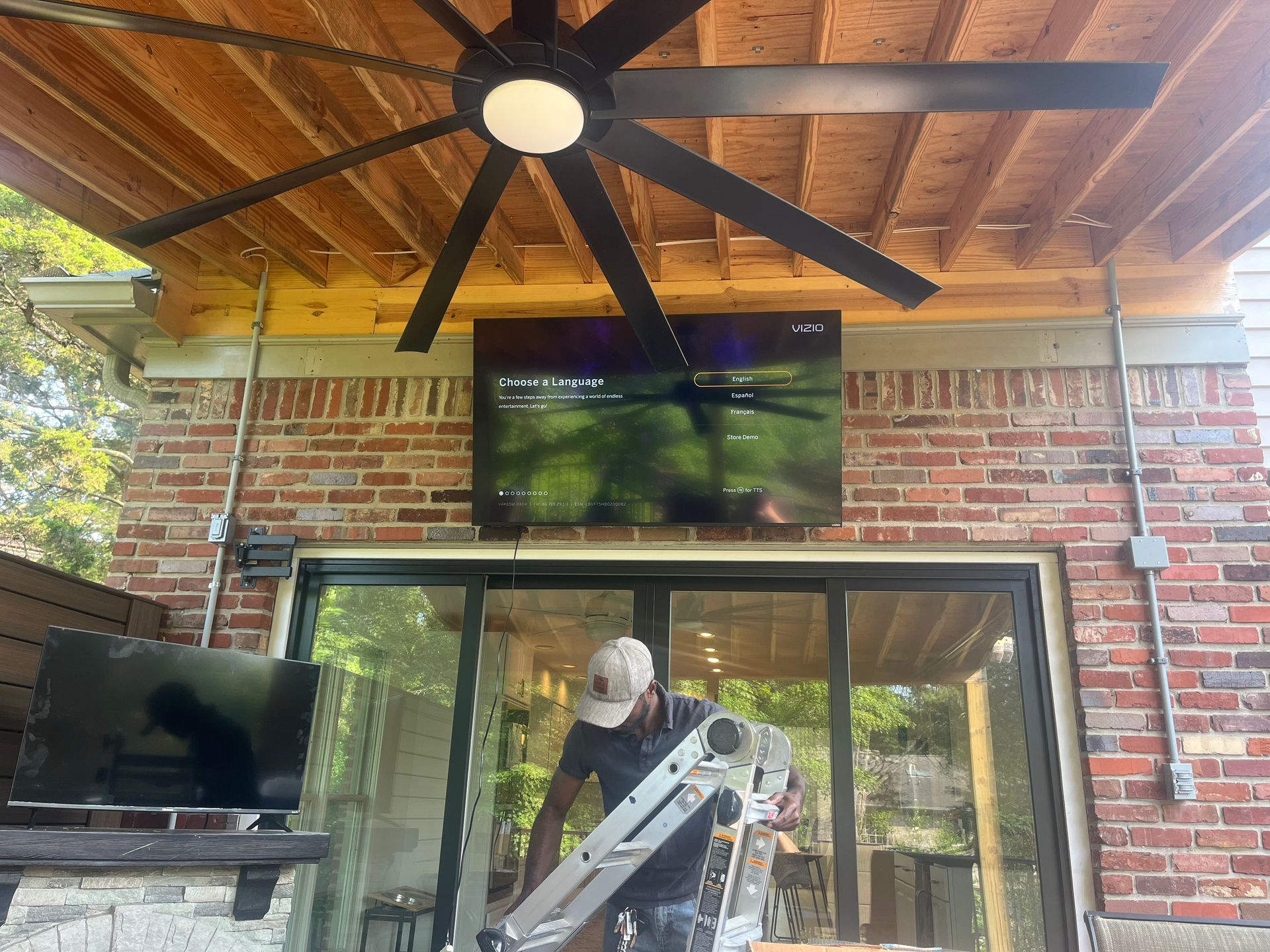 Person sets up ladder under outdoor TV. Large ceiling fan above. Brick wall with electrical conduits.