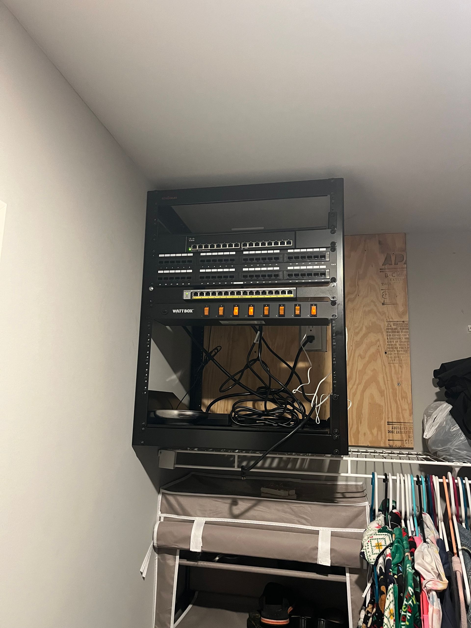 Black equipment rack mounted on a wall above a closet. Cables and electronic components are visible.