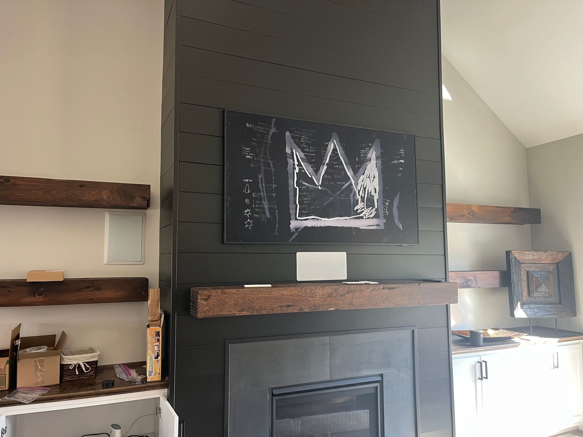 Fireplace with dark green shiplap, a Basquiat-style artwork, and a wooden beam mantel. Beige walls.