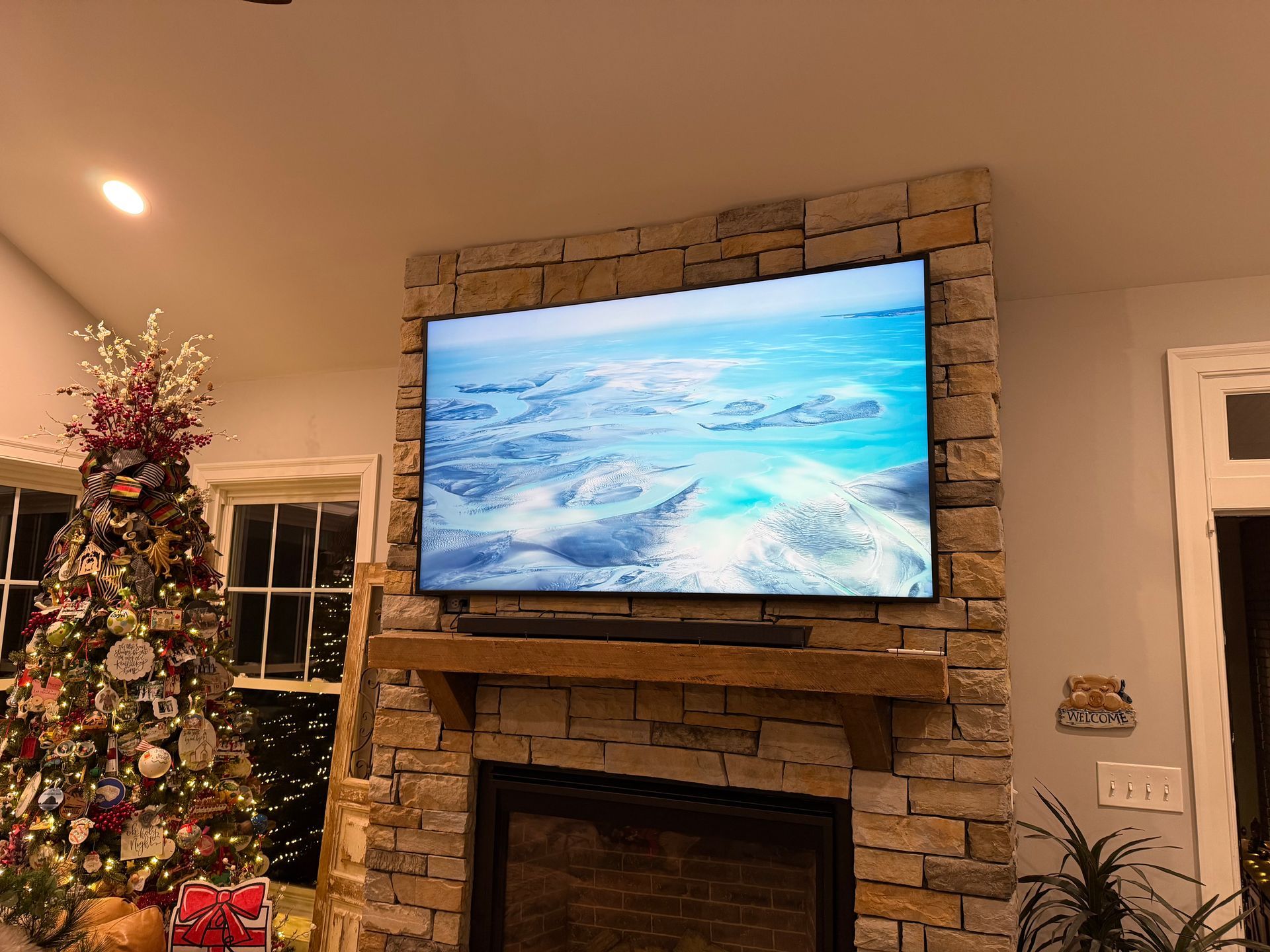 TV mounted above fireplace, Christmas tree in the corner, room with stone wall. 