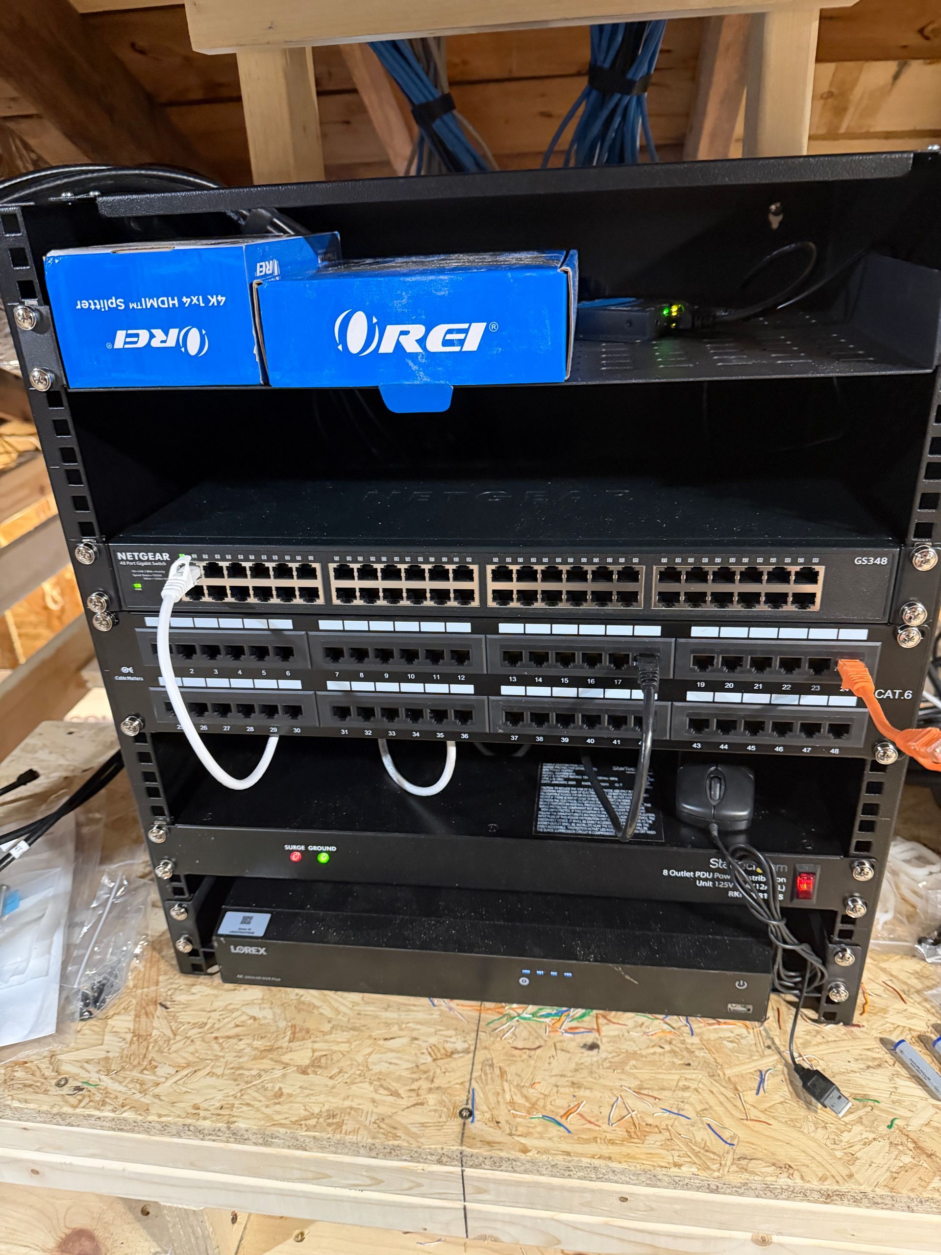 Black network rack in attic, holding network switch, patch panels, and other equipment. Blue cables above. 