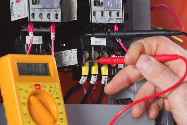 A person uses a multimeter to test electrical wiring in a panel. Wires are black, red, and yellow.