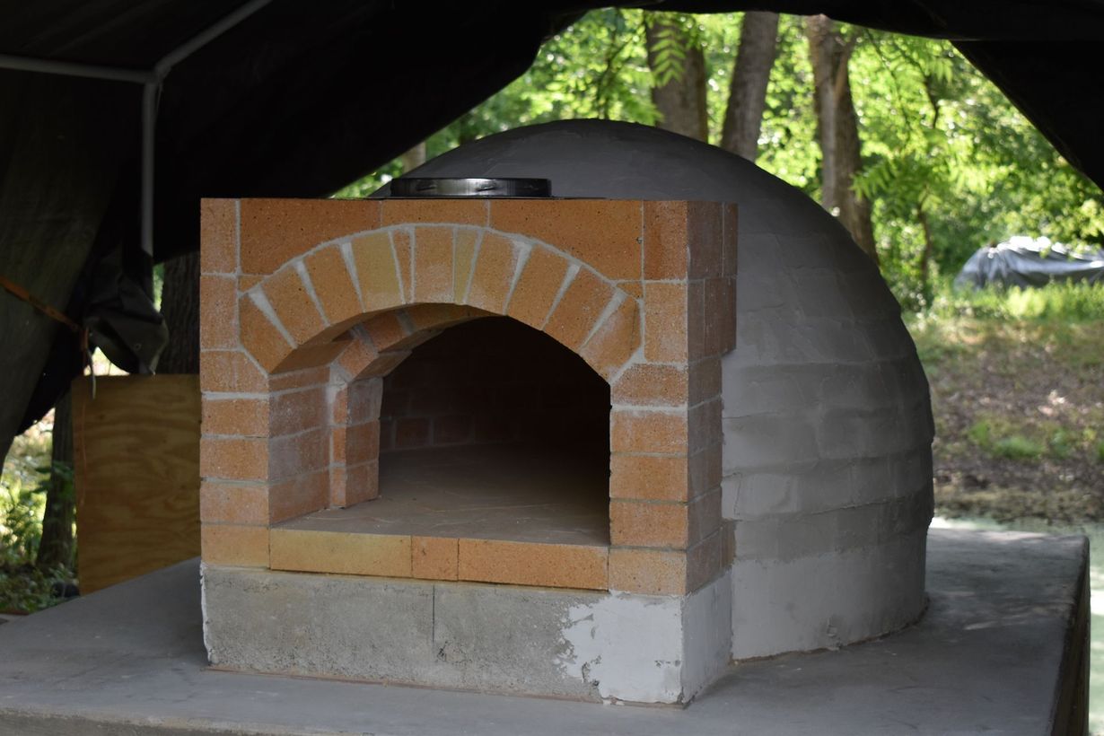 Brick outdoor wood-fired pizza oven on a concrete slab under a canopy, with trees in the background