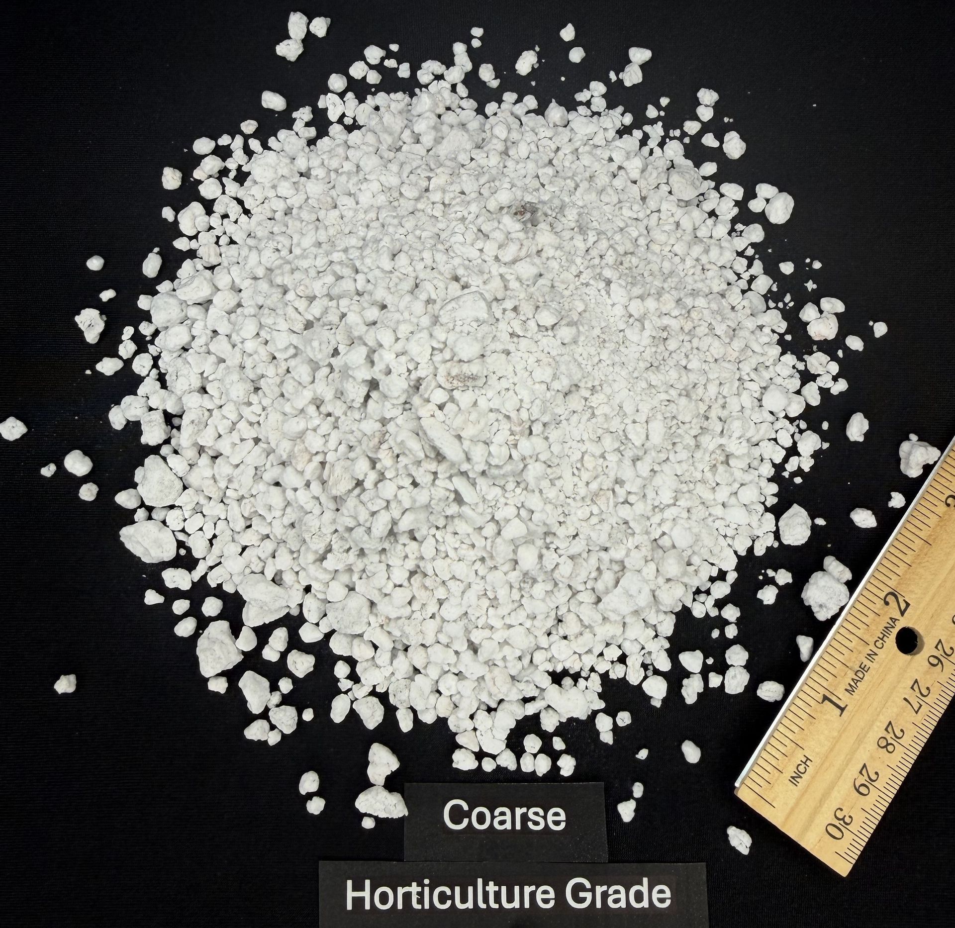 Pile of white coarse horticulture-grade granules beside a ruler on a black background