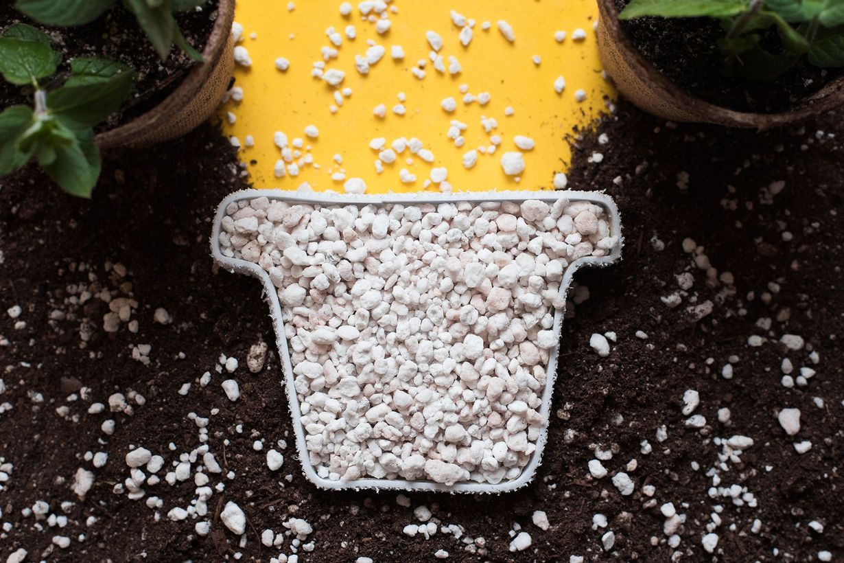 Yellow watering can pouring white pebbles into a white pot on dark soil, with potted plants nearby
