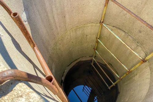 Looking down a narrow rusted ladder into a deep concrete well with blue water at the bottom
