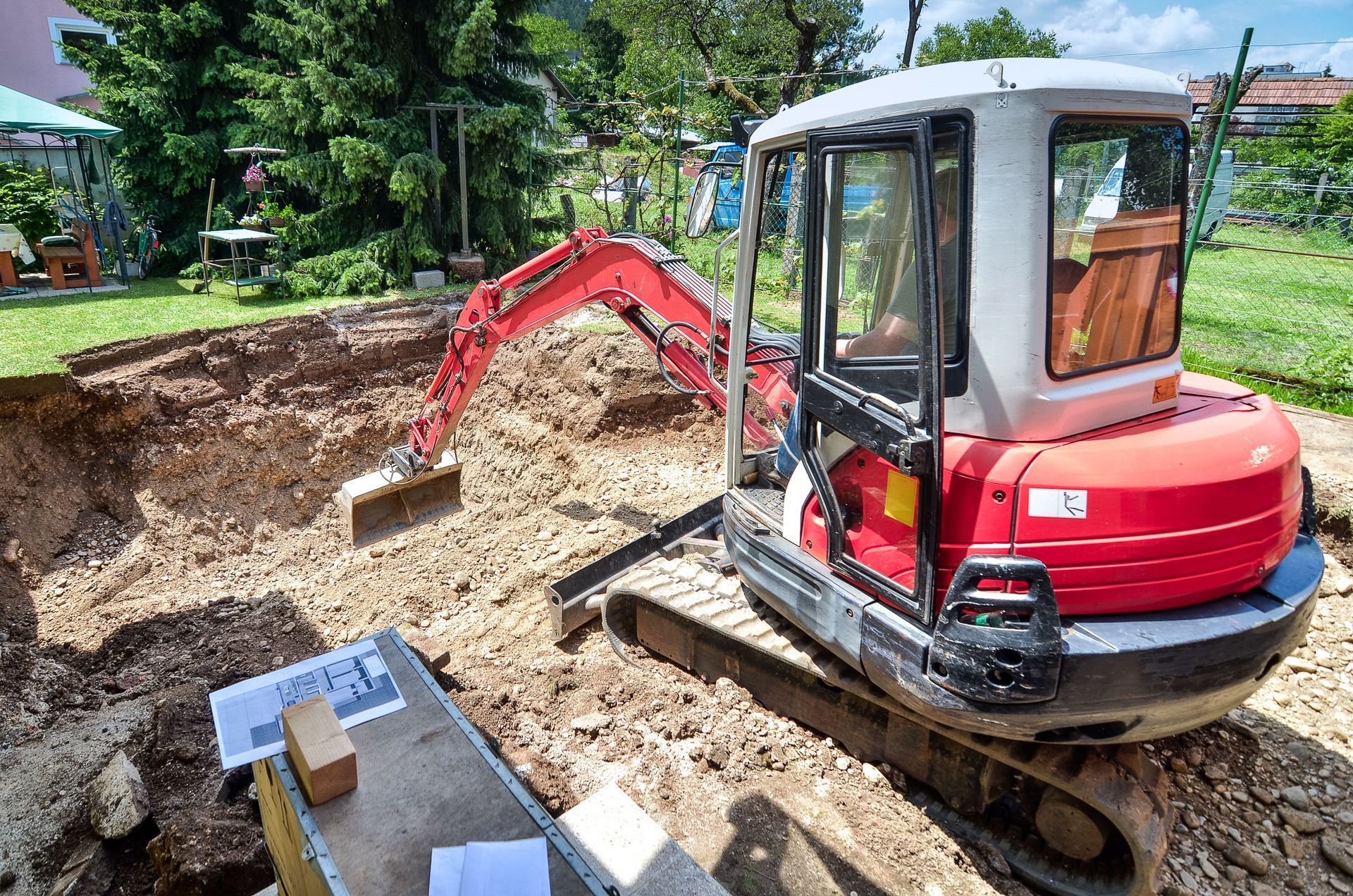 Red mini excavator digging in a muddy yard beside a gravel pile and cardboard boxes.