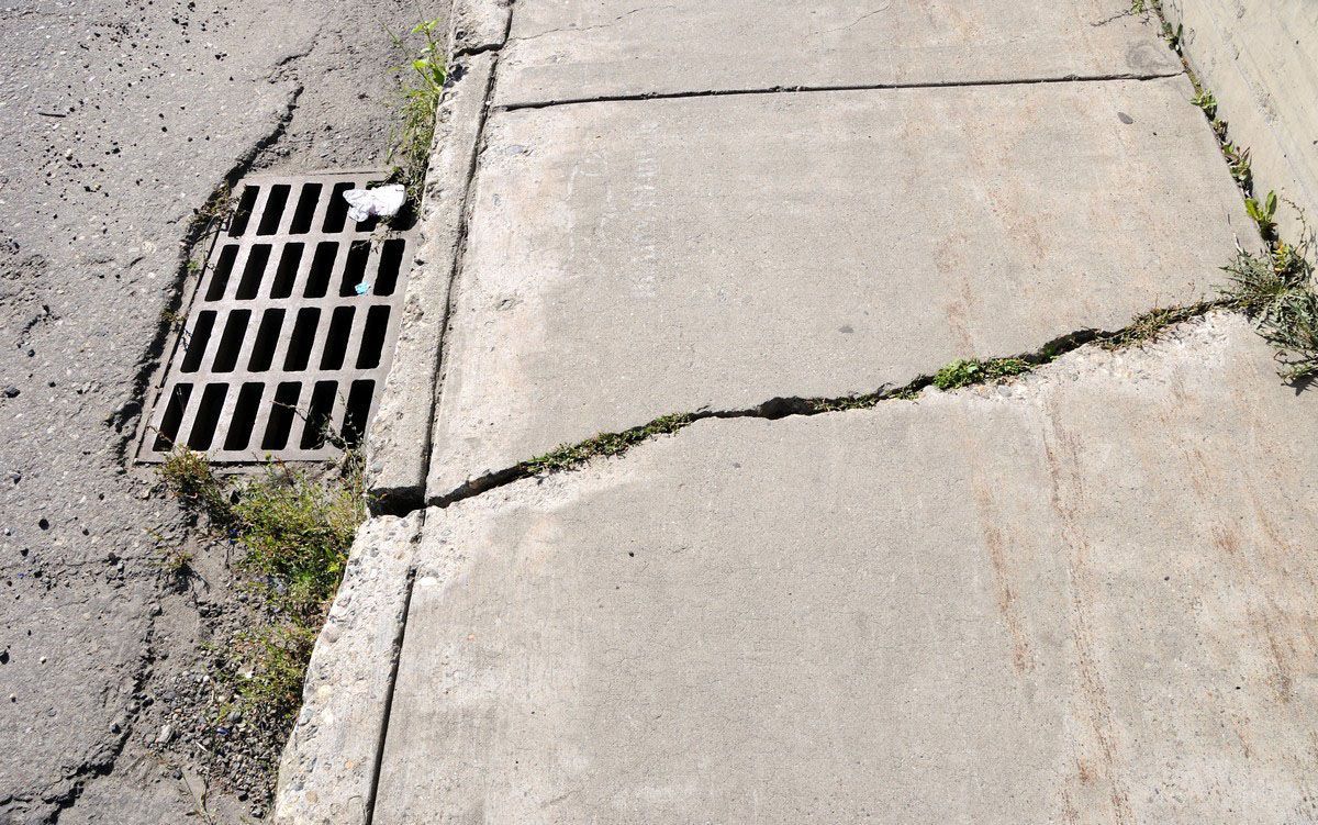 Cracked sidewalk beside a street drain grate with weeds growing in the gaps