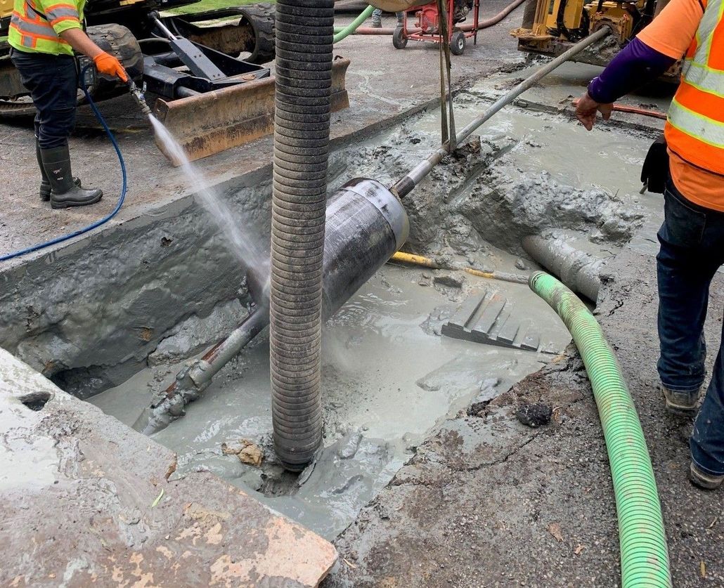 Workers hose down wet concrete around a buried pipe at a construction site.