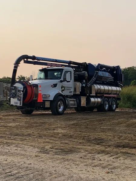 White vacuum truck with red hoses parked on a dirt field at sunset.