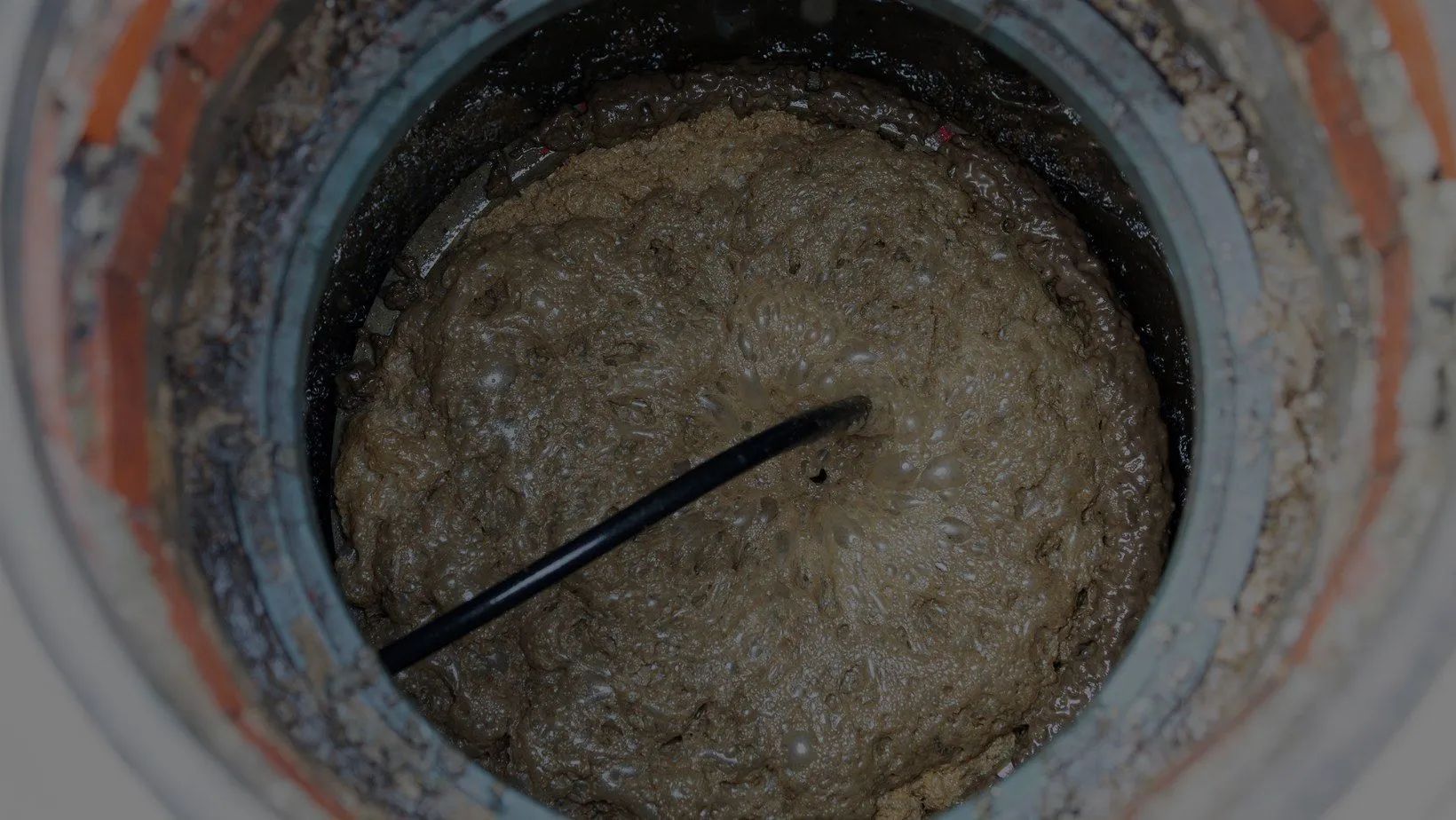 Top view into a circular pipe filled with murky water and sediment, with a black rod crossing it