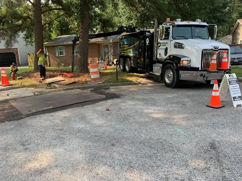 Tow truck parked at roadside construction site with cones, excavator, and a damaged utility area near a house