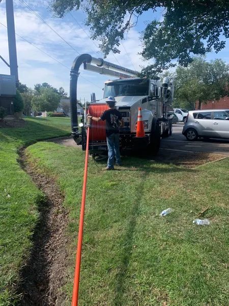 Worker by a red utility truck using a large vacuum hose on a grassy roadside.